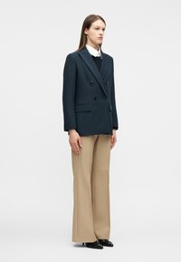 Γυναίκα που στέκεται φορώντας ένα διπλόμπουκλωτό μπλε σακάκι, λευκό πουκάμισο με γιακά, μπεζ παντελόνι φαρδύ στο πόδι και μαύρα παπούτσια με μυτερή μύτη.