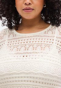 Εκρού πλεκτό τοπ με ανοιχτό σχέδιο που συνδυάζει ποικίλες υφές και μοτίβα, περιλαμβάνοντας οριζόντιες και κάθετες γραμμές.