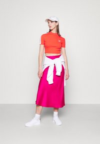 Cropped shirt rossa con dettagli bianchi, gonna midi in satin fucsia, sneaker bianche e cappellino da baseball bianco, in piedi su uno sfondo grigio chiaro.