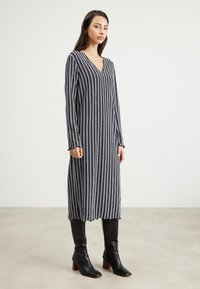 Robe longue à col en V avec des rayures verticales bleu marine et blanches, en matière côtelée. Dotée de manches longues et d'un ourlet volanté. Portée avec des bottes noires jusqu'au genou.