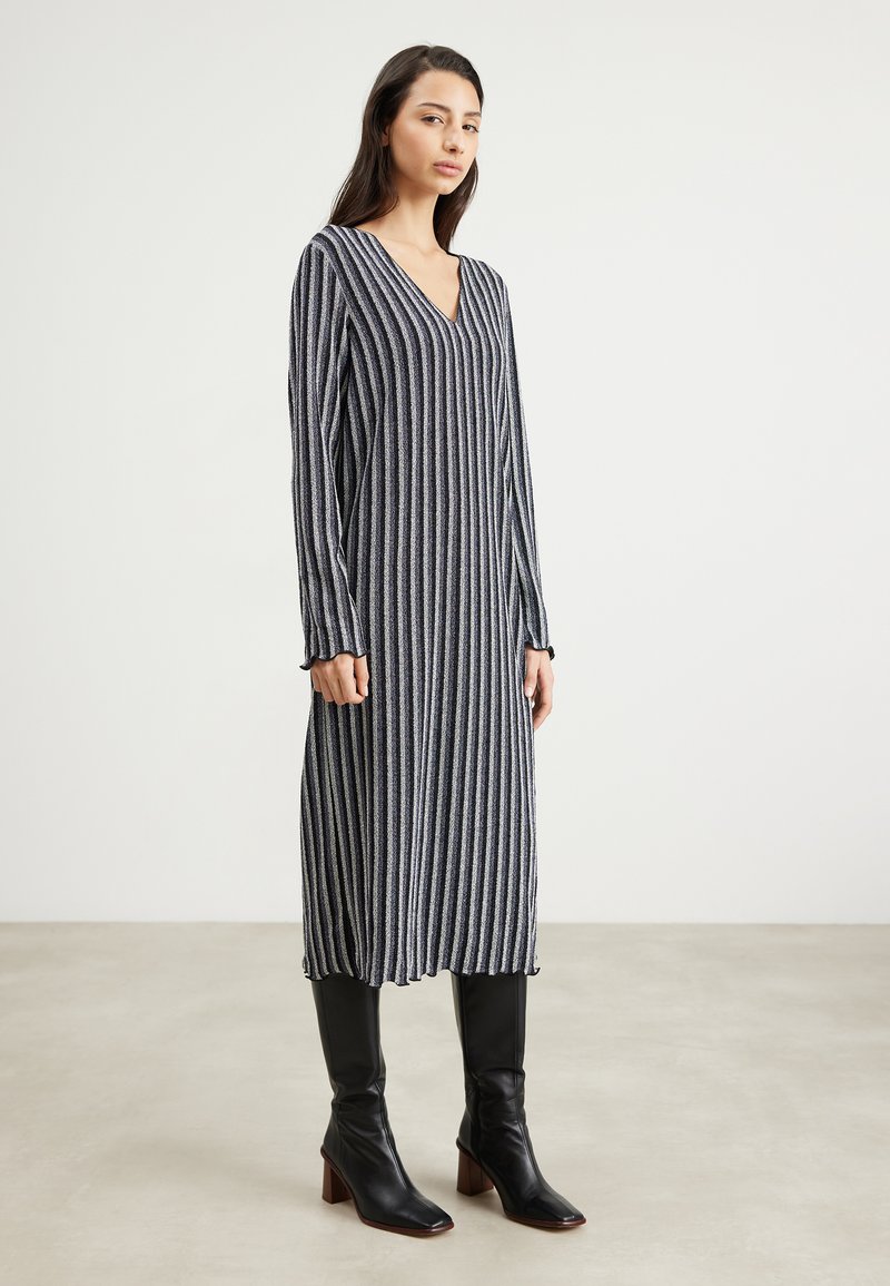Robe longue à col en V avec des rayures verticales bleu marine et blanches, en matière côtelée. Dotée de manches longues et d'un ourlet volanté. Portée avec des bottes noires jusqu'au genou.