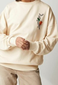 Bluza w kolorze kremowym o luźnym kroju, z haftowanym kwiatowym wzorem z pomarańczowymi i zielonymi akcentami po lewej stronie.