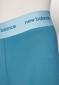 Artimu atstumu matomos mėlyni sportiniai legginai su šviesiai mėlynu juosmeniu, kuriame tamsiai mėlyna spalva užrašyta "new balance" logotipas.
