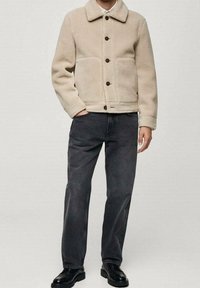 Beige Fleecejacke mit Kragen, vorderem Knopfverschluss und zwei großen Taschen, kombiniert mit dunkelgrauen Jeans und schwarzen Schuhen.