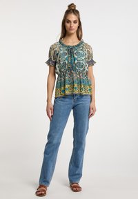 Top a maniche corte con motivo floreale in blu e giallo, con scollatura arricciata e maniche a volantino, abbinato a jeans blu chiaro e sandali marroni.