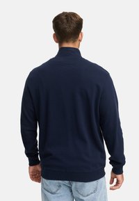 Hombre con un suéter de manga larga azul marino y jeans claros, visto desde atrás sobre un fondo claro y liso.