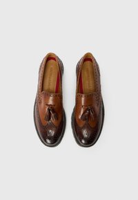 Brūni ādas slip-on apavi ar wingtip dizainu, ar dekoratīvām perforācijām un pušķiem, iekšpuses odere ir sarkana.