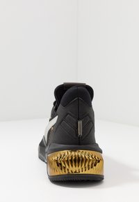 Schwarzer Sportschuh mit einem strukturierten goldenen Absatz, weißen seitlichen Akzenten und einer gepolsterten Silhouette. Entwickelt für Komfort und Leistung.