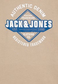 Tela beige con un gráfico central en azul y blanco, que muestra "Jack & Jones", con una forma de diamante y detalles de texto.