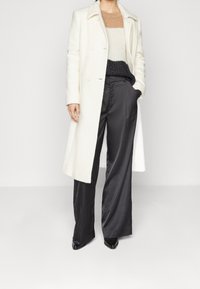 Manteau en laine blanche, pull en tricot noir et beige, pantalon large en satin noir, chaussures noires brillantes. Design minimaliste avec une coupe structurée.