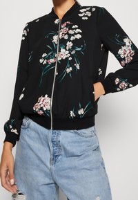 Czarna kurtka bomber z kwiatowym wzorem w bieli i turkusie, wyposażona w zamek błyskawiczny, ściągacze na mankietach i pasie. Lekka tkanina.