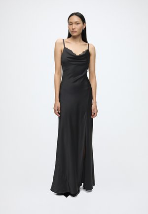 Femme portant une longue robe noire en satin avec des bretelles fines et des détails en dentelle, debout devant un fond blanc uni.
