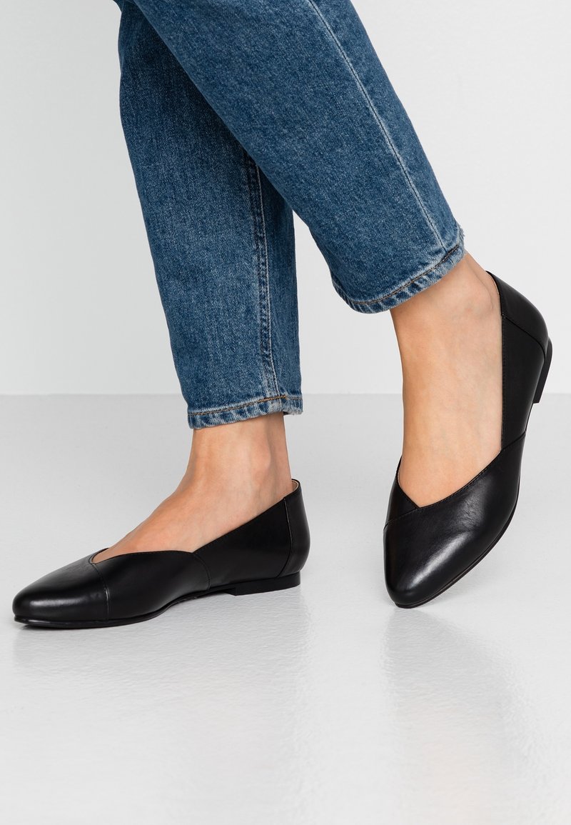 Bailarinas de cuero negro con punta afilada, con un recorte en forma de V, tacón bajo y textura suave. Combinadas con jeans de mezclilla cortos.