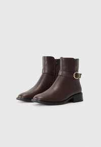 Botas de tobillo de cuero marrón con puntera redondeada, cremallera lateral, tacón apilado y una correa decorativa con un acento circular dorado.
