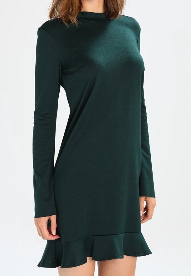 Femme portant une robe verte foncée à manches longues avec un col montant et un ourlet à volants, debout devant un fond clair uni.