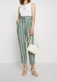 Pantalones de pierna ancha a rayas verdes con cintura ajustable, combinados con una camiseta sin mangas blanca y un bolso de mimbre blanco. Sandalias de tacón blanco con punta abierta.