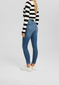 Mujer vestida con jeans ajustados de tiro alto en azul, suéter corto a rayas en blanco y negro, y zapatillas blancas, de pie frente a un fondo liso.