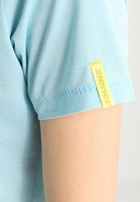 Svetle modrá košile s krátkým rukávem vyrobená z texturované tkaniny, s žlutou cedulkou s nápisem "Ziener" na levém rukávu. Úhledné šití podél lemování.