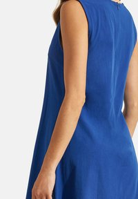 Robe sans manches bleue avec une texture lisse, une taille ajustée et une fermeture éclair dissimulée au dos. Son design simple souligne l'élégance.