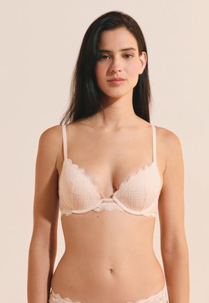 Joven mujer con cabello largo y oscuro que lleva un sujetador de encaje beige claro y ropa interior a juego sobre un fondo liso.
