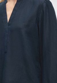 Gros plan d'une personne portant une chemise à manches longues bleu foncé avec un col en V et une couture visible le long de l'encolure.