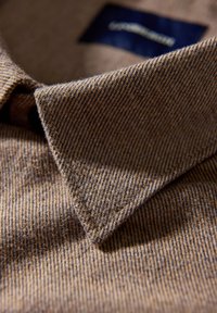 Bruine geweven stof met een visgraatpatroon en een kraag. Bevat een donkerblauw label dat gedeeltelijk zichtbaar is binnen de kraag.