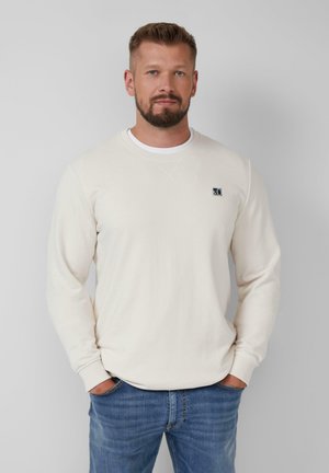 Sweatshirt crème clair à col rond avec des manches longues et un petit logo cousu sur la poitrine. Associé à un jean bleu délavé. Texture lisse.
