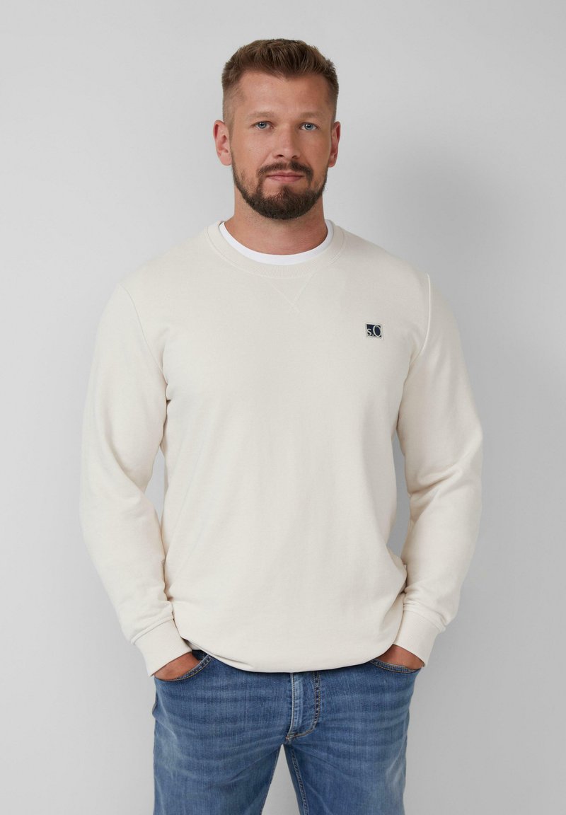 Sweatshirt crème clair à col rond avec des manches longues et un petit logo cousu sur la poitrine. Associé à un jean bleu délavé. Texture lisse.