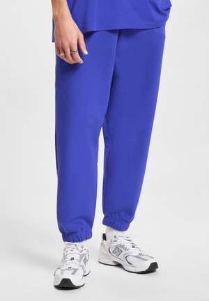 Blue cotton joggers with elastic cuffs, featuring side pockets, paired with white and gray athletic sneakers and a loose-fitting top.