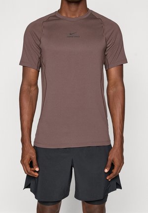 Atlet iført brun Nike Pro kortærmet trøje og sorte atletikshorts, stående mod en ensfarvet lys baggrund.