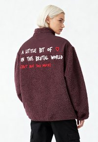 Veste en polaire bordeaux avec un col montant, comportant un texte blanc et rouge au dos. Texture douce avec une coupe décontractée et des manches longues.