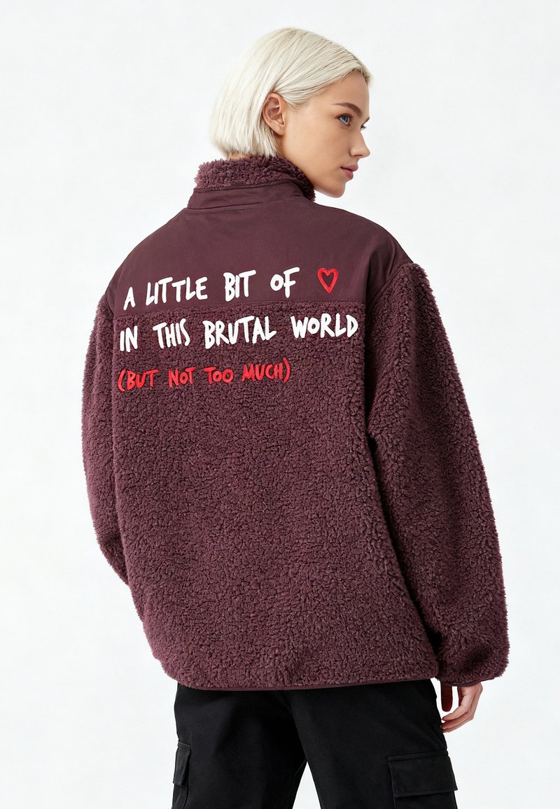 Veste en polaire bordeaux avec un col montant, comportant un texte blanc et rouge au dos. Texture douce avec une coupe décontractée et des manches longues.