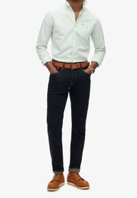 Lichtgroen overhemd met knopen, donkerblauwe spijkerbroek, bruine leren riem en beige veterlaarzen. Het overhemd heeft een zak en omgeslagen manchetten.