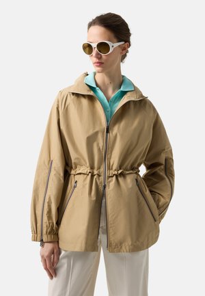 Kvinde iført beige lynlåstrøje med indsnøret talje, lyseblå skjorte med krave, hvide bukser og store hvide solbriller, kigger mod venstre.