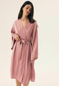 Różowy satynowy szlafrok z szerokimi rękawami, zapinany na zakładkę, z dopasowanym paskiem. Gładka tekstura, długość do kolan oraz boczne rozcięcia.