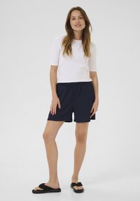Haut à manches courtes blanc et shorts navy à taille élastique. Modèle portant des tongs noires. Design simple, motifs minimaux, texture lisse.