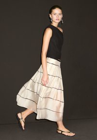 Femme marchant dans un haut noir sans manches, une jupe crème fluide avec de fines rayures noires et des sandales noires sur fond sombre.