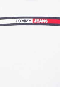 Camiseta de algodón blanca con un diseño de rayas horizontales en azul marino y rojo, con "TOMMY" en azul marino y "JEANS" en blanco sobre un fondo rojo.