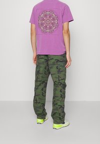 Violetti puuvillainen t-paita, jossa on värikäs mandalakuosi selässä, yhdistettynä vihreisiin maastokuvioisiin cargo-housuihin ja kirkkaan keltaisiin tennareihin.