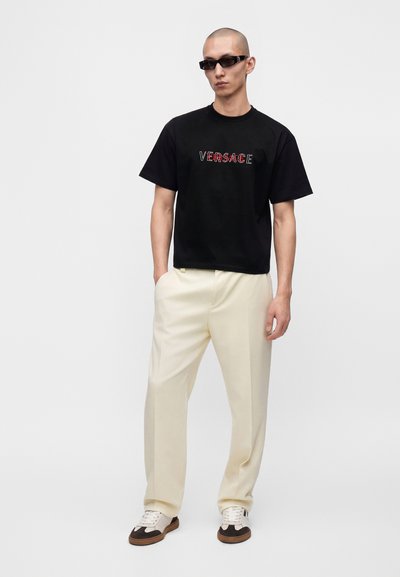 Άνδρας που φοράει μαύρο μπλουζάκι Versace, κρεμ παντελόνια, λευκά και καφέ αθλητικά παπούτσια και μαύρα γυαλιά ηλίου, στέκεται με τα χέρια στις τσέπες.