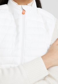 White padded vest with a high collar and a front zipper featuring an orange pull tab. The model wears a cream knit sweater under it.