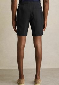 Pantaloni scurți negri cu croială dreaptă, cu brâie, un buzunar pe spate și o textură netedă. Purtați cu pantofi slip-on de culoare maro deschis.