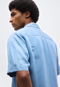 Νεαρός άνδρας με σγουρά μαλλιά που φοράει ανοιχτό μπλε πουκάμισο από τζιν με μικρά σχέδια μαύρης γραμμής στην πλάτη, σε απλή φόντο.