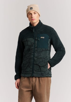 Joven con gorro beige, chaqueta polar texturizada verde oscuro con cremallera y bolsillo en el pecho, y pantalones de pana marrones, de pie con una mano en el bolsillo.
