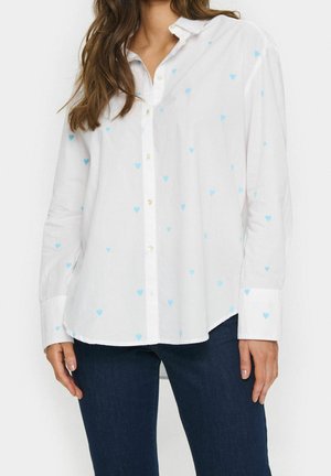 Vrouw die een wit overhemd draagt met lichtblauwe hartpatronen en donkerblauwe spijkerbroek, staand tegen een effen achtergrond.