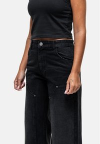 Pantaloni wide-leg in denim nero con tasche anteriori, chiusura con bottone e dettagli cuciti. Il tessuto ha una texture leggermente sbiadita.