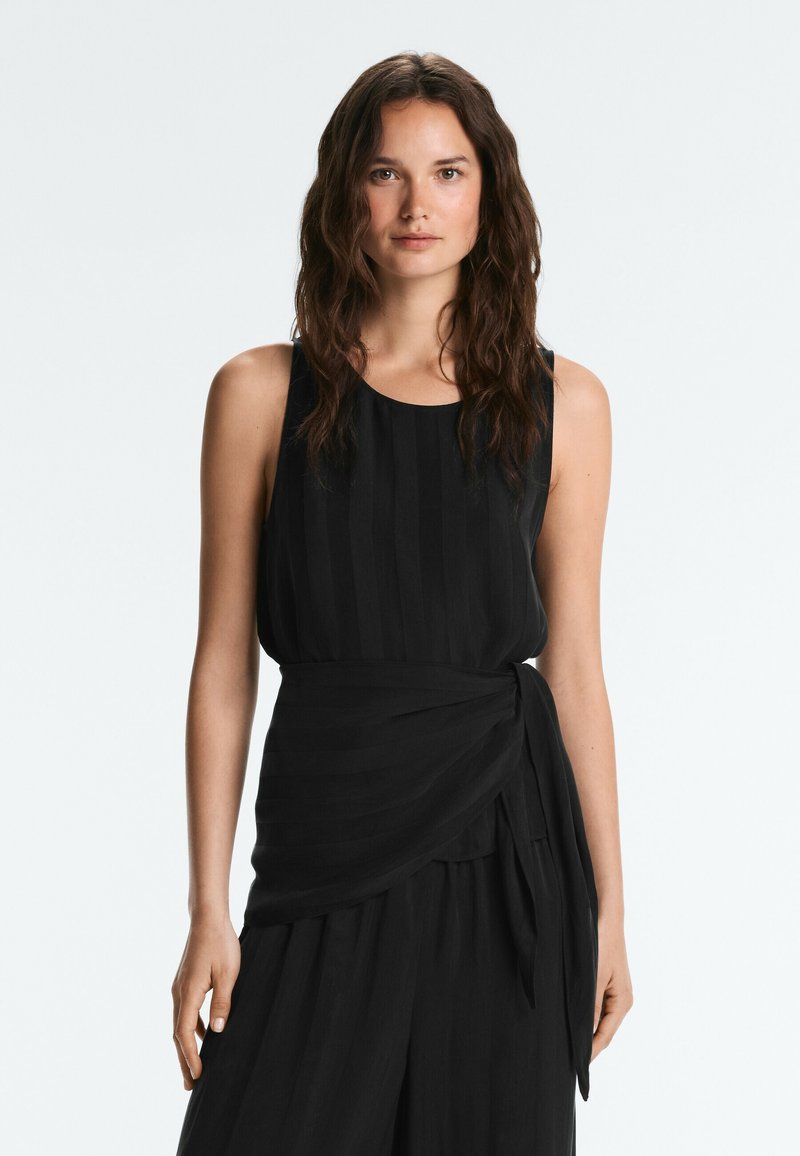 Femme aux longs cheveux bruns ondulés portant une robe noire sans manches avec ceinture à la taille, debout devant un fond clair uni, face à l'avant.