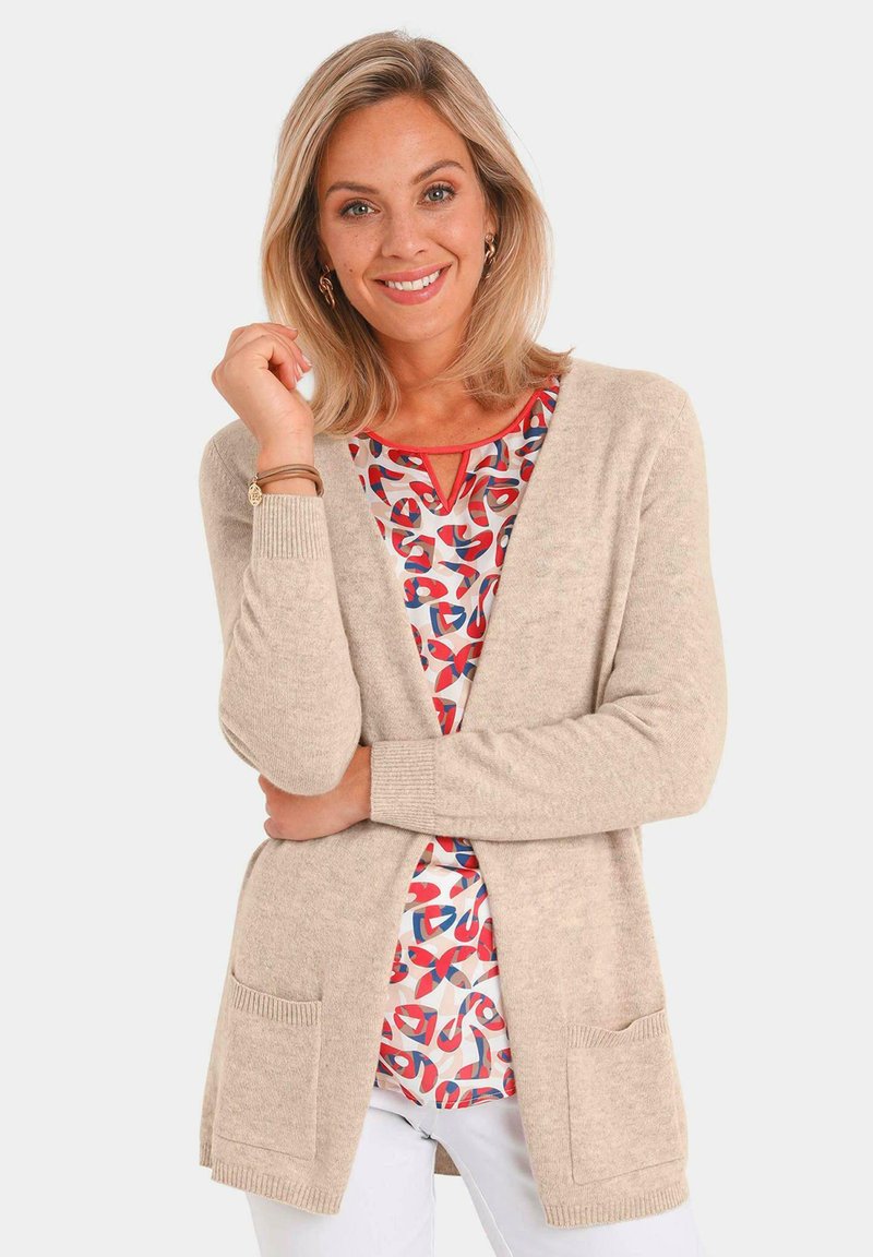 Cardigan beige à l'avant ouvert, avec des poignets côtelés et deux poches avant, porté avec un haut coloré à motif abstrait en dessous.