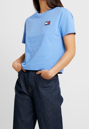 Nainen, joka pukeutuu vaaleansiniseen cropattu t-paitaan, jossa on pieni Tommy Jeans -logo, ja korkeavyötäröisiin tummansinisihin farkkuihin, kädet taskuissa.