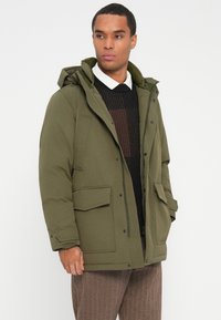 Férfi olajzöld kapucnis parkát visel fekete pulóver és fehér galléros ing felett, barna mintás nadrággal párosítva, fehér háttér előtt áll.